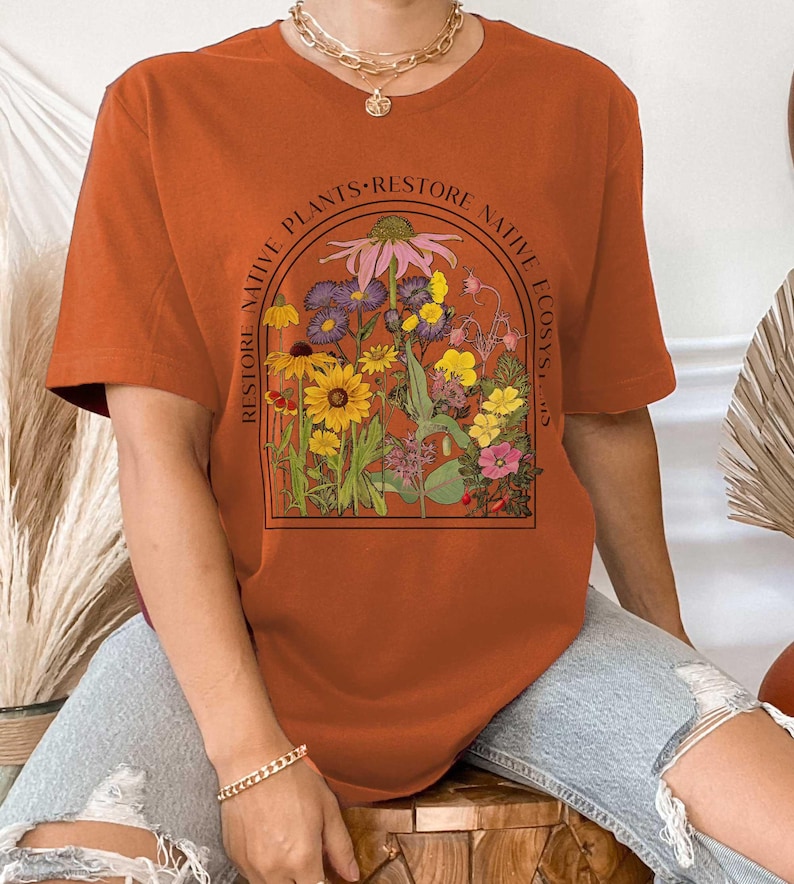 K&ouml;nnte beinhalten: Ein rostfarbenes T-Shirt mit einem grafischen Druck einer Vielzahl von Blumen in einem rechteckigen Rahmen. Der Text "RESTORE NATIVE PLANTS RESTORE NATIVE ECOSYSTEMS" ist um den Rahmen herum gedruckt.