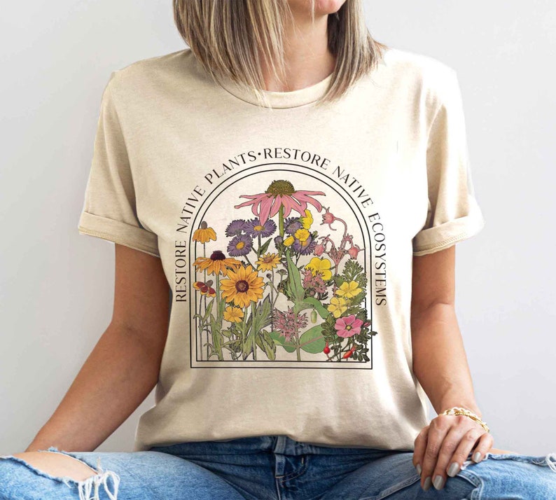 K&ouml;nnte beinhalten: Ein beiges T-Shirt mit einer Grafik aus verschiedenen Blumen in einem runden Rahmen. Der Text "Restore Native Plants. Restore Native Ecosystems" umgibt den Rahmen.