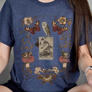 May include: A navy blue t-shirt with a vintage illustration of a barn owl, butterflies, frogs, and a skeleton. The illustration is surrounded by floral vines and mushrooms.