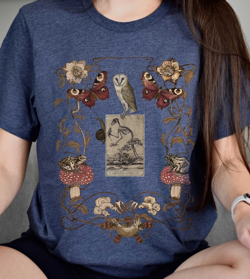 May include: A navy blue t-shirt with a vintage illustration of a barn owl, butterflies, frogs, and a skeleton. The illustration is surrounded by floral vines and mushrooms.