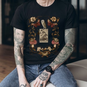 May include: Black t-shirt with a vintage illustration of a bird, butterflies, frogs, and mushrooms. The illustration is surrounded by a floral border.