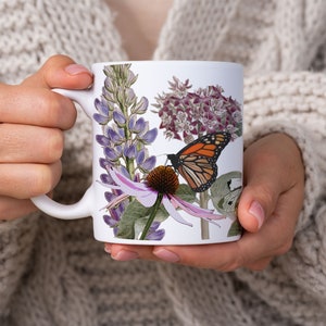 May include: White ceramic mug with a floral design featuring purple lupine flowers, a pink coneflower, and a monarch butterfly. The butterfly is orange, black, and white.