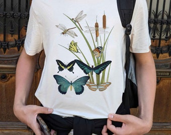 May include: White t-shirt featuring a detailed illustration of various butterflies and dragonflies among reeds and grasses. The design includes butterflies in shades of blue, yellow, and brown. The shirt is made of a soft, light-colored fabric.