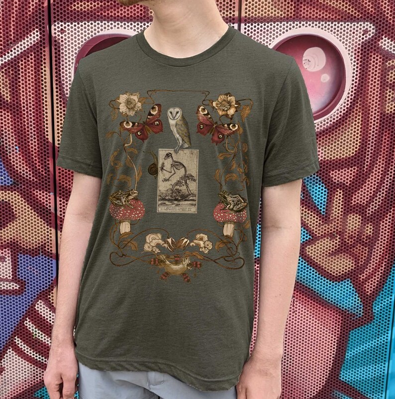 May include: A green t-shirt with a vintage illustration of an owl, butterflies, frogs, and mushrooms. The illustration is surrounded by a decorative floral border.