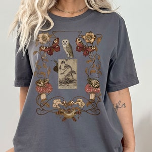 May include: A dark gray t-shirt with a vintage-style print featuring a skeleton, owls, butterflies, frogs, and mushrooms. The print is surrounded by a decorative floral border.