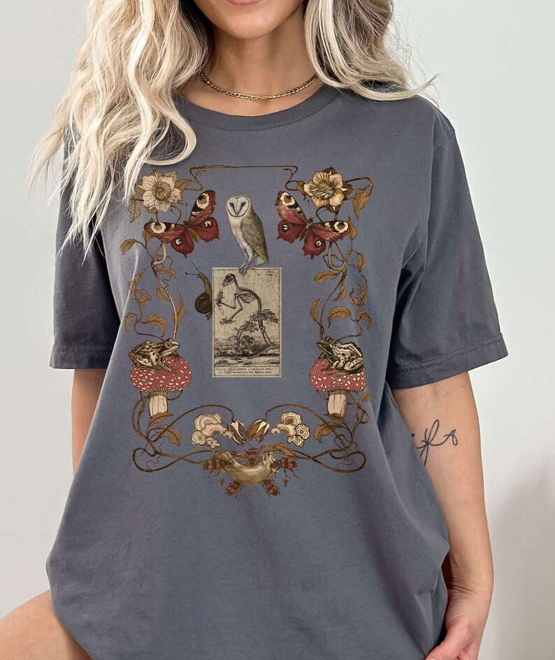 May include: A dark gray t-shirt with a vintage-style print featuring a skeleton, owls, butterflies, frogs, and mushrooms. The print is surrounded by a decorative floral border.