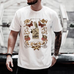 May include: White t-shirt with a vintage illustration of an owl, mushrooms, butterflies, and flowers. The illustration is framed with a decorative border.