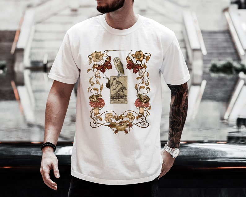 May include: White t-shirt with a vintage illustration of an owl, mushrooms, butterflies, and flowers. The illustration is framed with a decorative border.