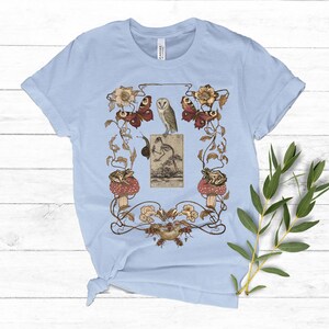 May include: Light blue t-shirt with a vintage illustration of an owl, butterflies, frogs, and mushrooms in a floral design.