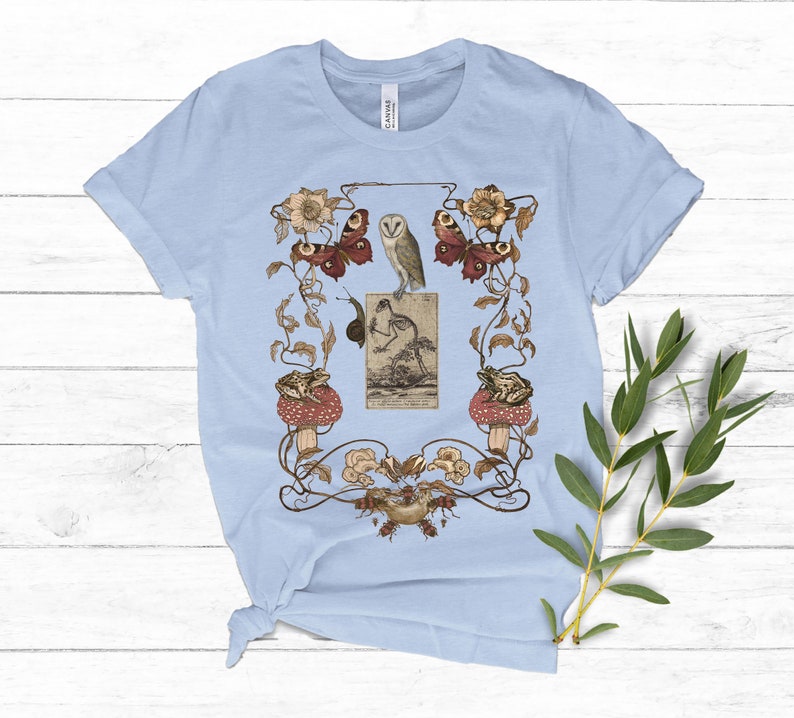 May include: Light blue t-shirt with a vintage illustration of an owl, butterflies, frogs, and mushrooms in a floral design.