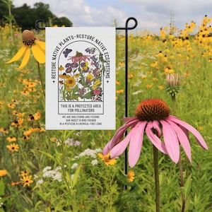 May include: A white garden flag with a colourful illustration of wildflowers and the text "Restore Native Plants - Restore Native Ecosystems". The text below the illustration reads "This is a protected area for pollinators. We are sheltering and feeding our insect & bird friends in a pesticide & chemical-free zone."