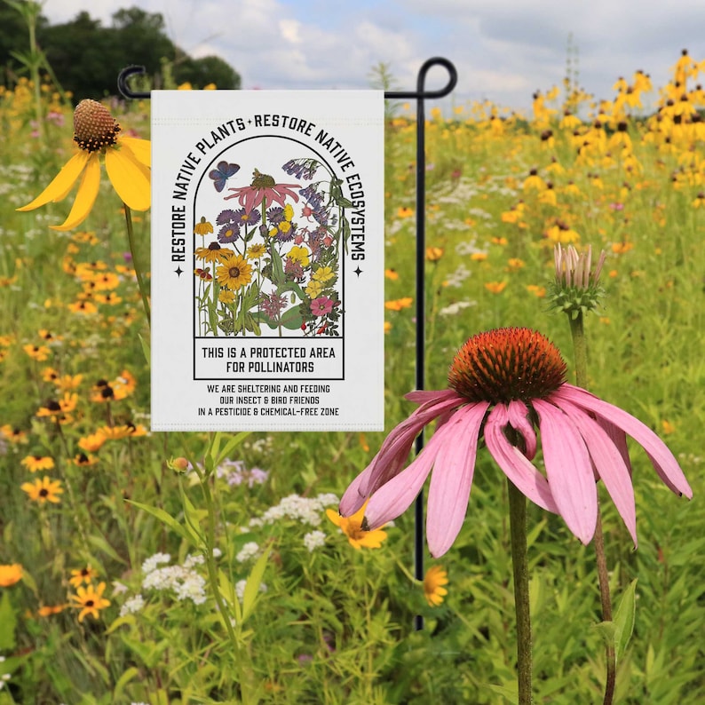 May include: A white garden flag with a colourful illustration of wildflowers and the text "Restore Native Plants - Restore Native Ecosystems". The text below the illustration reads "This is a protected area for pollinators. We are sheltering and feeding our insect & bird friends in a pesticide & chemical-free zone."
