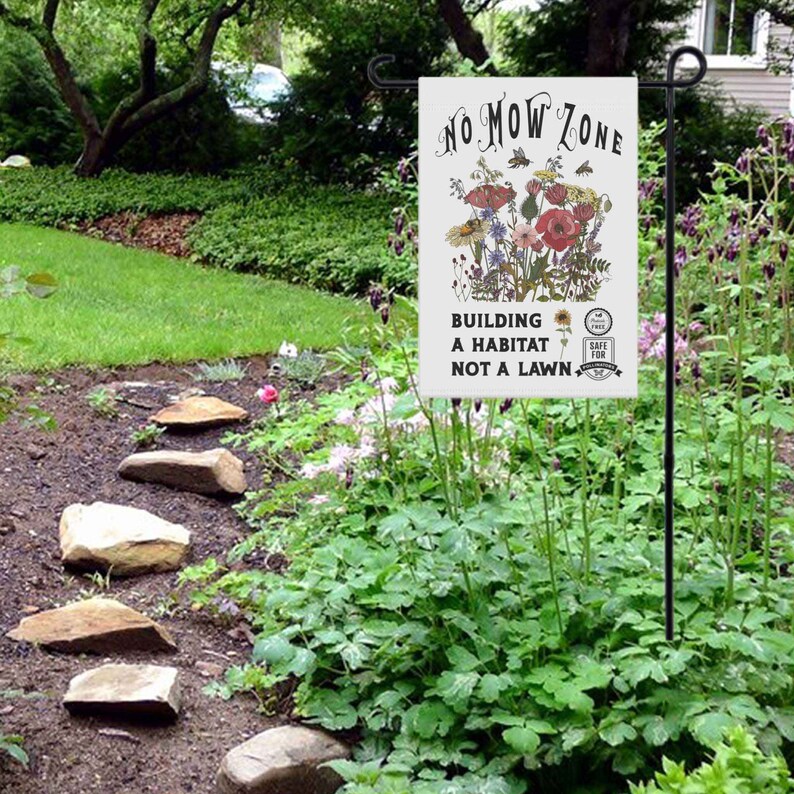K&ouml;nnte beinhalten: Eine wei&szlig;e Gartenflagge mit dem Text "NO MOW ZONE" und einer floralen Illustration. Die Flagge tr&auml;gt auch die Aufschrift "BUILDING A HABITAT NOT A LAWN" und enth&auml;lt ein "SAFE FOR BEES" Emblem. Die Flagge wird in einem Garten gezeigt.