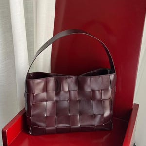 Puede incluir: Un bolso de mano de cuero color burdeos profundo con un patrón de rejilla trenzada en el exterior. El bolso tiene un asa arqueada única unida a la parte superior, una base rectangular y un acabado semibrillante en las tiras de cuero trenzadas.