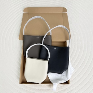 May include: Three structured handbags in cream, charcoal grey, and black, each featuring a single top handle wrapped in white protective paper. The bags are arranged inside an open cardboard shipping box, with the brand name The Row visible.