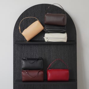 Puede incluir: Una colección de seis bolsos cilíndricos en varios colores, incluyendo beige, marrón oscuro, negro, blanco y rojo. Cada bolso cuenta con una correa fina, un cierre de cremallera superior y un acabado liso, exhibidos en dos estantes de color negro.