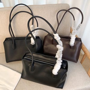 May include: Three rectangular shoulder bags in black and dark brown. Each bag features a top zipper closure and two long handles, with one handle on each bag wrapped in protective white tissue paper. The bags have a smooth, solid-colored finish.