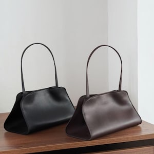May include: Two structured handbags with a smooth finish and a single thin handle. One handbag is solid black, and the other is solid dark brown. Both bags feature a wide, open-ended base design with curved side panels and a minimalist silhouette.
