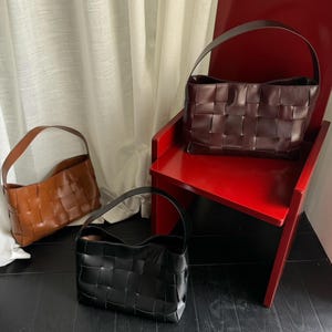 May include: Three woven handbags in different colors. One large dark brown bag sits on a red chair, while a medium tan bag and a medium black bag are positioned nearby. Each bag features a wide, interlaced leather-like strap and body design.
