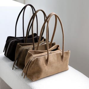 Può includere: Tre borse a spalla rettangolari disposte in fila, caratterizzate da una morbida consistenza in camoscio. Le borse sono nei colori beige, verde oliva e marrone scuro. Ogni borsa presenta una chiusura superiore con zip e due manici lunghi.