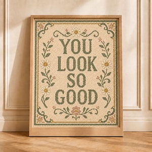 May include: A framed wall art piece featuring the text YOU LOOK SO GOOD in dark green, composed of small circular elements. The design includes a decorative border of green vines and white daisy flowers with yellow centers on a cream background.
