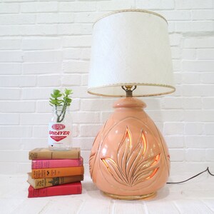 May include: A peach-colored ceramic table lamp with a white lampshade. The lamp base has a carved design of leaves and features a warm glow when lit.