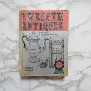 May include: A vintage book titled "Twelfth Antiques and their Current Prices" by Edwin G. Warman. The cover features a black and white illustration of a teapot and a clock. The book is a 25th anniversary edition.