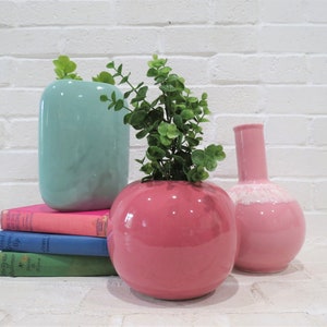 May include: Three ceramic vases in different shapes and colors. A light blue vase with a rounded rectangular shape, a pink vase with a round shape, and a pink vase with a round body and a tall, thin neck. The round pink vase has a white rim around the top. The vases are arranged on a stack of colorful books.