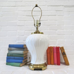 May include: White ceramic table lamp with a gold base and a ribbed design. The lamp is on a white brick wall background with a stack of books in front of it.