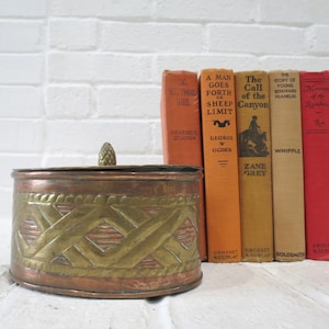 Peut inclure: Une boîte à bijoux ronde en cuivre et en laiton avec un motif tissé décoratif. La boîte a un couvercle avec un petit fleuron sur le dessus. La boîte est placée devant une pile de livres anciens.
