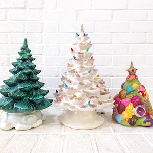 Puede incluir: Tres figuras de árbol de Navidad de cerámica. Una es verde con una base blanca, otra es blanca con purpurina y una base blanca, y la última es verde con adornos coloridos y una base blanca.