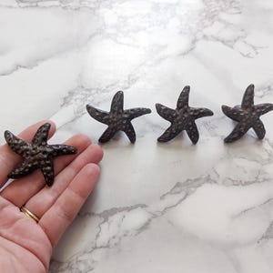 May include: Four bronze-coloured starfish decorations. One starfish is held in a hand, while the other three are arranged on a white and grey marble-patterned surface. The starfish have a textured surface and a five-point design.