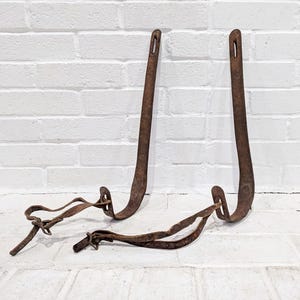 May include: Two antique, rusty brown metal hooks with a curved design and a small hole at the top. Two additional rusty metal pieces with a looped design are also present. The items are displayed against a white brick wall.