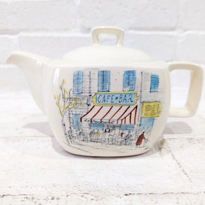 Może przedstawiać: Biały ceramiczny czajnik z namalowanym na boku obrazkiem przedstawiającym kawiarnię. Scena przedstawia kawiarnię z pasiastym daszkiem i napisem "Cafe-Bar" nad daszkiem. Przed kawiarnią siedzą ludzie.