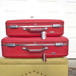 May include: Two stacked red hard-shell suitcases with silver metal latches and handles. Each suitcase features a small, dangling luggage tag with red, white, and blue stripes. The bottom suitcase has a small silver rectangular brand label.