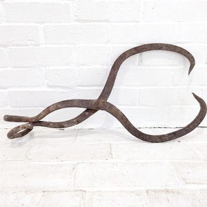 May include: An antique, rusty metal tool with curved arms and a looped handle. The tool is likely used for handling hot objects. The metal has a weathered appearance, suggesting age and use. The tool is set against a white brick wall.