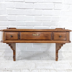 May include: A vintage wooden wall-mounted shelf with three drawers and decorative brass hardware. The shelf is a warm brown color and features ornate detailing on the front and supporting brackets. The drawers have round knobs.
