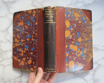 Könnte beinhalten: Ein Vintage-Buch mit dem Titel "Woman's Share in Primitive Culture" von Otis Tufton Mason. Das Buch hat einen marmorierten Einband mit roten, orangefarbenen und blauen Wirbeln. Das Buch ist geöffnet und der Rücken ist sichtbar.
