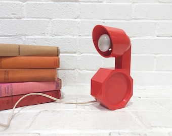 Könnte beinhalten: Eine rote Tischlampe aus Kunststoff mit einem runden Sockel und einem flexiblen Arm. Die Lampe hat eine weiße Glühbirne und ein weißes Kabel.