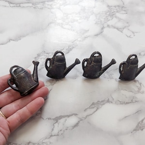 May include: Four small, bronze-colored watering can figurines. One is held in a hand, while the other three are arranged on a white and gray marble surface. The watering cans have a handle, spout, and a small opening for filling.