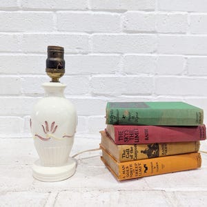 May include: A cream-colored ceramic table lamp with a brown socket and a decorative floral design. The lamp is next to a stack of vintage books with colorful covers. The books include titles like "The Sky's the Limit" and "A Man Goes Forth".