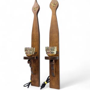 May include: Two wooden wall sconces with a dark brown finish. Each sconce features a decorative gold-colored eagle emblem at the top and a small shelf with a gold-colored candle holder. Black electrical cords are visible.