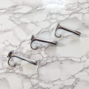 Op de afbeelding: Drie zilverkleurige metalen haken met een gebogen ontwerp, geplaatst op een wit en grijs marmerpatroon oppervlak. De haken hebben een eenvoudig, functioneel ontwerp, geschikt voor het ophangen van items. Geschikt voor interieurdecoratie.