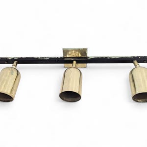 Op de afbeelding: Een vintage armatuur met drie lampen, kegelvormige tinten in messingkleur en een zwarte balk. De armatuur heeft een vierkante montageplaat en staat tegen een witte achtergrond. De tinten zijn gelijkmatig verdeeld.