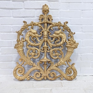 Puede incluir: Decoración mural dorada y ornamentada con un diseño simétrico. La pieza presenta patrones de volutas, figuras de animales estilizadas y un elemento vertical central. La forma general es un escudo o cresta estilizada, adecuada para la decoración de interiores.