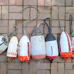 May include: A collection of seven weathered nautical buoys featuring various cylindrical and tapered shapes. These buoys are painted in white, red, and orange, with visible signs of wear, chipped paint, and attached rope loops for hanging.