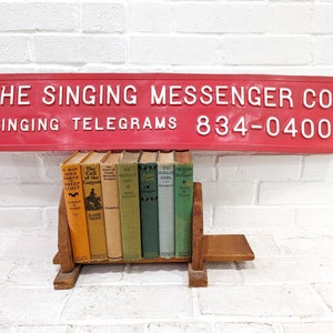 Könnte beinhalten: Ein rotes Schild mit weißen Buchstaben, auf dem "THE SINGING MESSENGER CO. SINGING TELEGRAMS 834-0400" steht. Ein hölzerner Buchstütze hält eine Reihe von Vintage-Taschenbüchern.