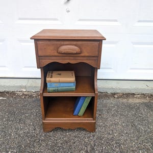 May include: A medium-brown wooden nightstand featuring a single top drawer with a curved handle, an open middle shelf holding three stacked books, and an open bottom shelf containing two upright books, all set against a white garage door.