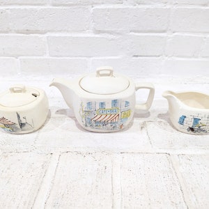 May include: A white ceramic tea set with a Parisian cafe scene design. The set includes a teapot, creamer, and sugar bowl.