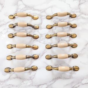 May include: A collection of twelve cabinet pulls with a cream-colored center and antique brass-toned metal ends. The pulls are arranged on a white and gray marble-patterned surface, showcasing their design and potential for home decor projects.
